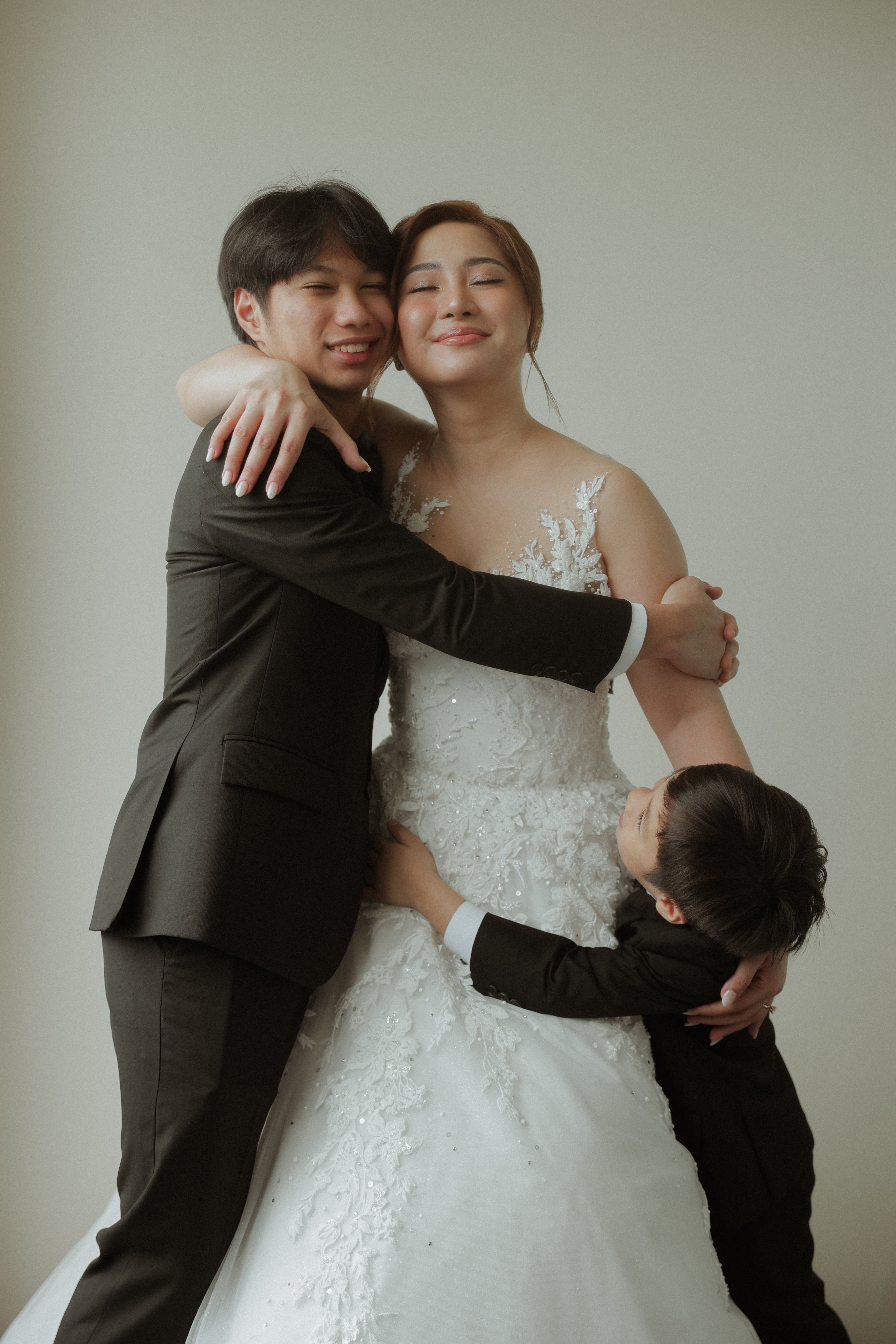 Man in a suit embracing a woman in a wedding dress with a child on her lap against a plain background