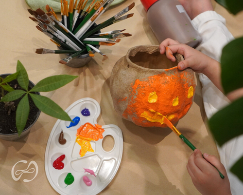 Person painting a ceramic pot with orange and yellow colors on a table with paintbrushes and a palette.